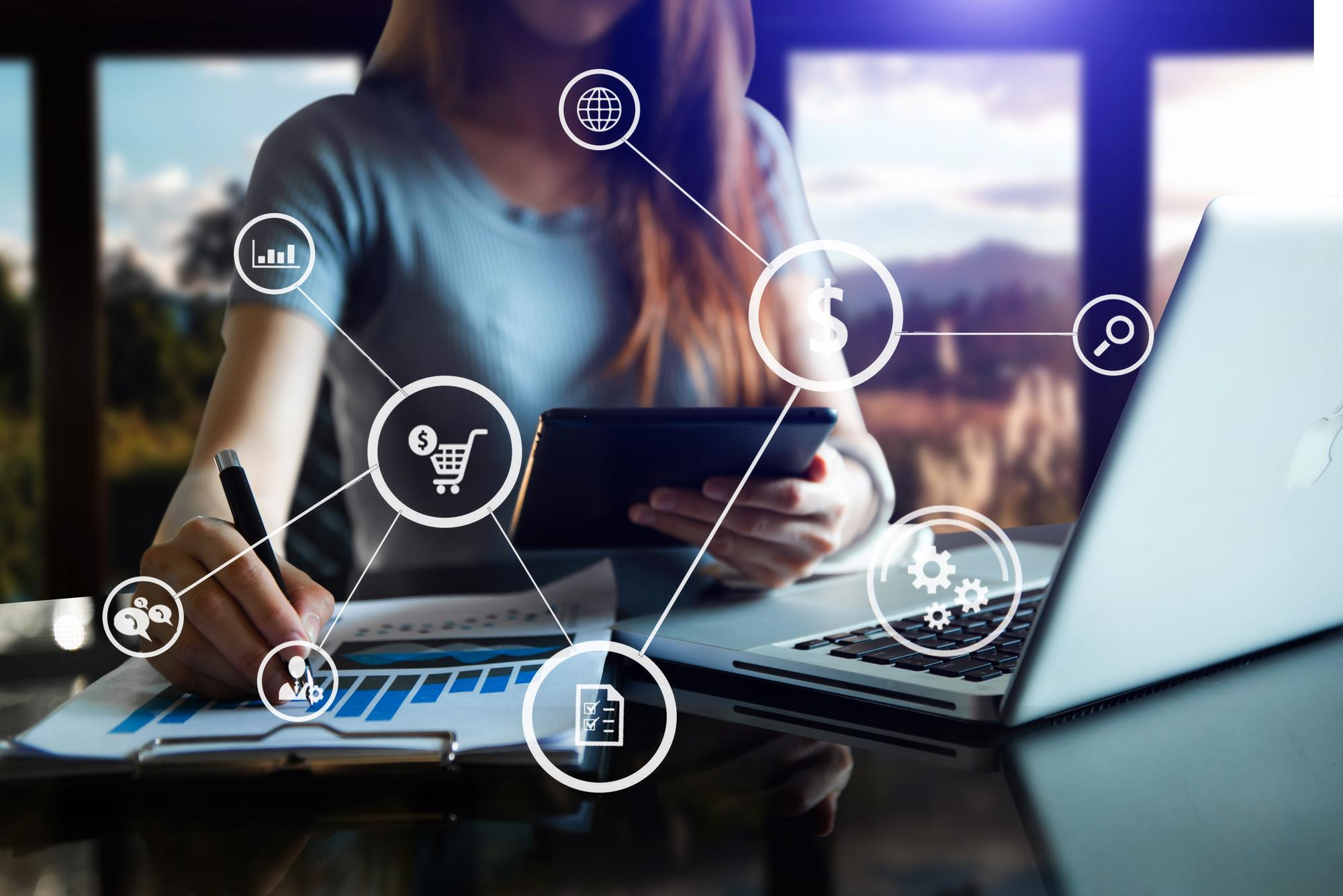 A marketer working at a laptop and tablet with a digital network graphic showing connected icons for e-commerce, sales revenue, SEO analytics, and customer communication.