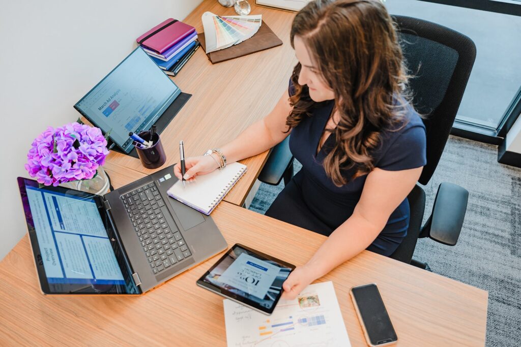 A female digital marketing strategist taking notes at a wooden desk with multiple laptops showing ad analytics, a tablet, and brand color swatches.